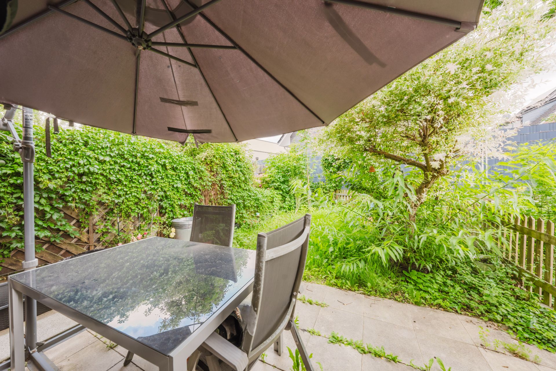 Terrasse mit Glastisch, Stühlen und großem Sonnenschirm neben grünem Garten.