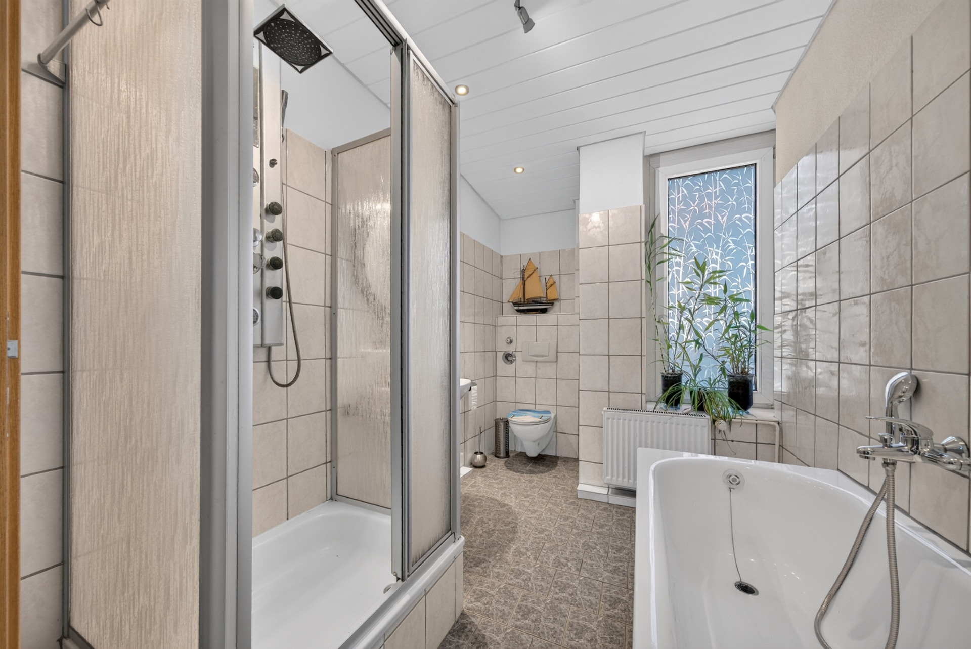 Helles Badezimmer mit Dusche, Badewanne, WC, Fenster und Pflanze auf der Fensterbank.