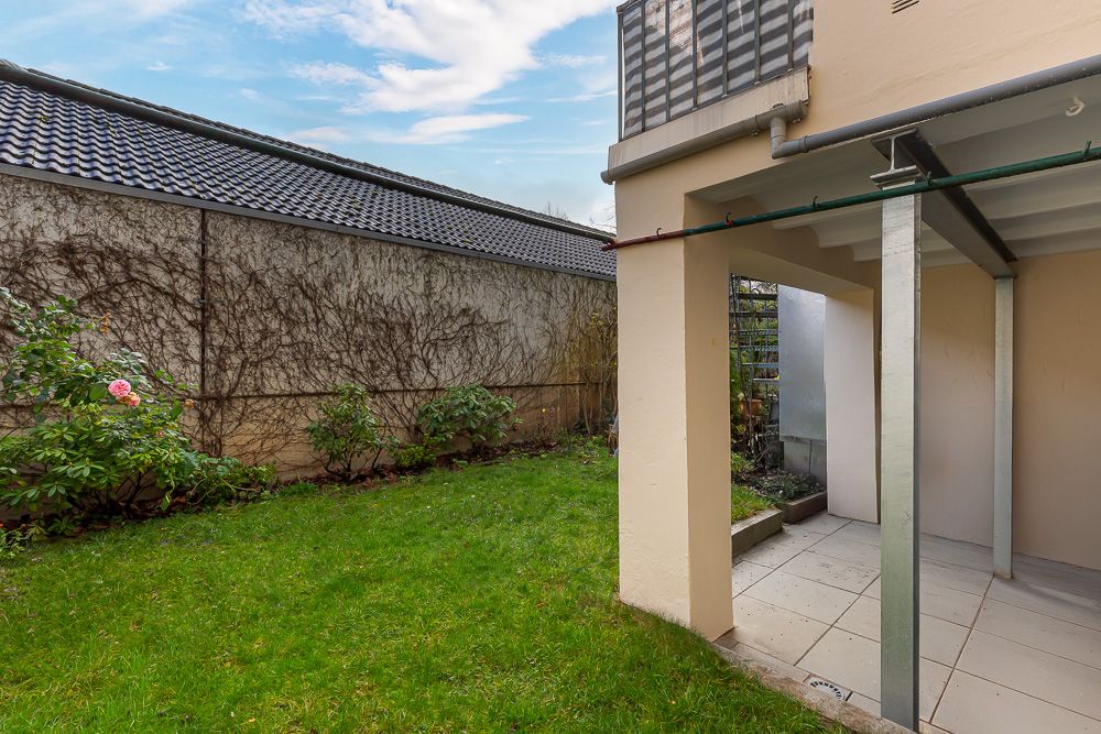 Kleiner Garten mit Rasen, Blumenbeeten und überdachter Terrasse neben einem Haus.