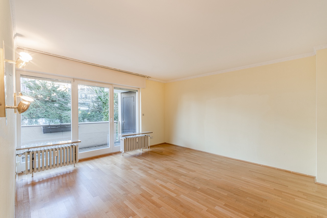 Helles, leeres Zimmer mit Parkettboden, großer Fensterfront und Zugang zu einem Balkon.