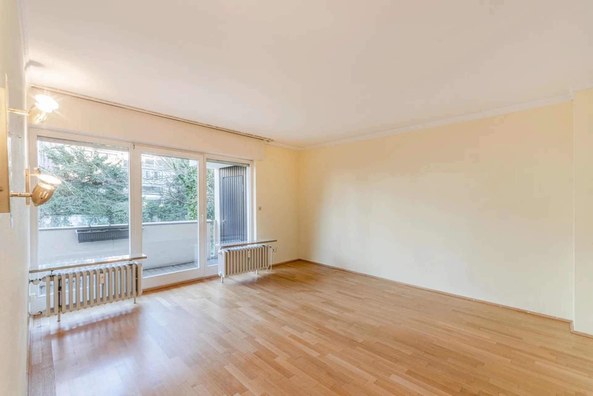 Helles, leeres Zimmer mit Parkettboden, großer Fensterfront und Zugang zu einem Balkon.