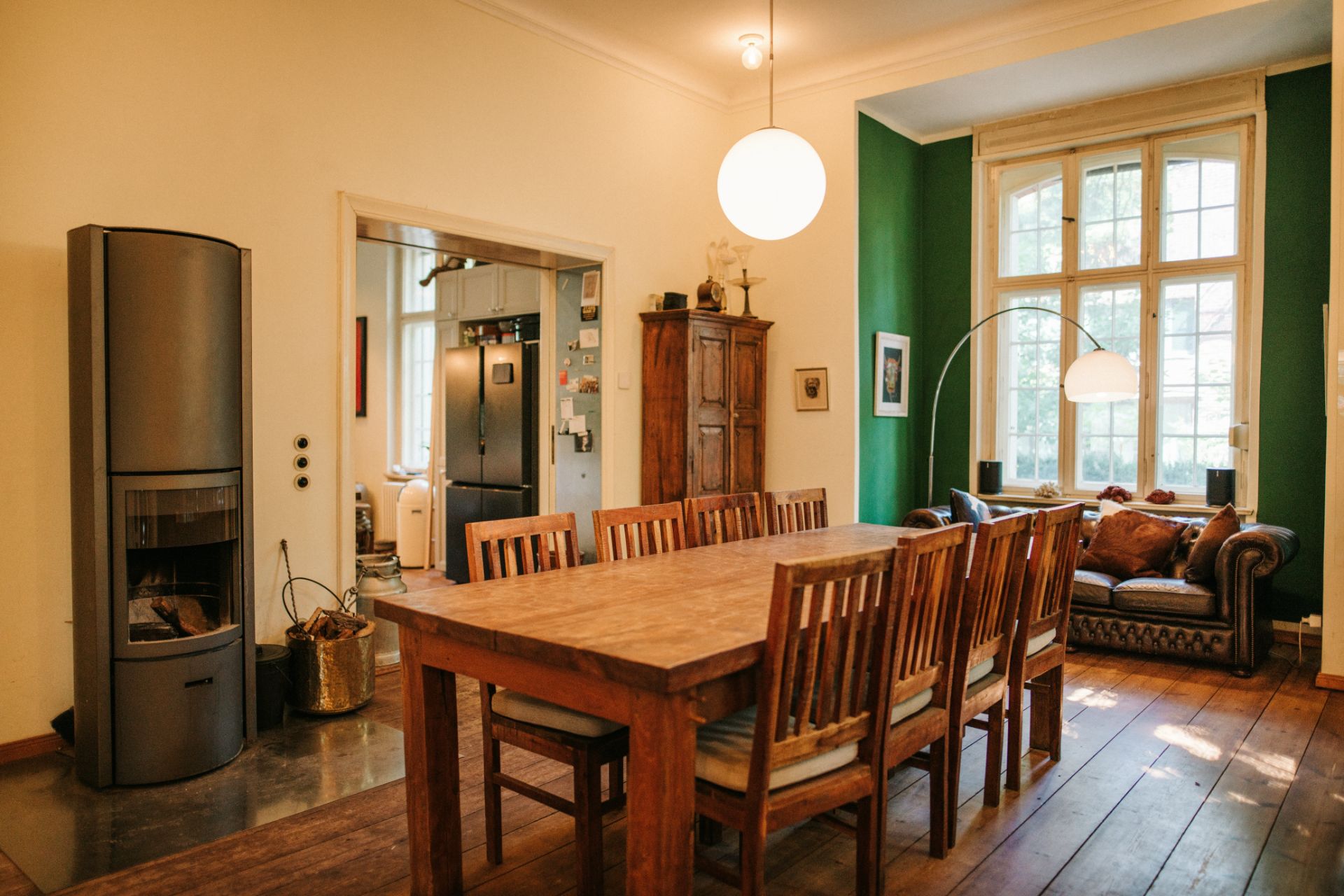 Esszimmer mit Holztisch, Stühlen, Kaminofen und Sofa vor großem Fenster.