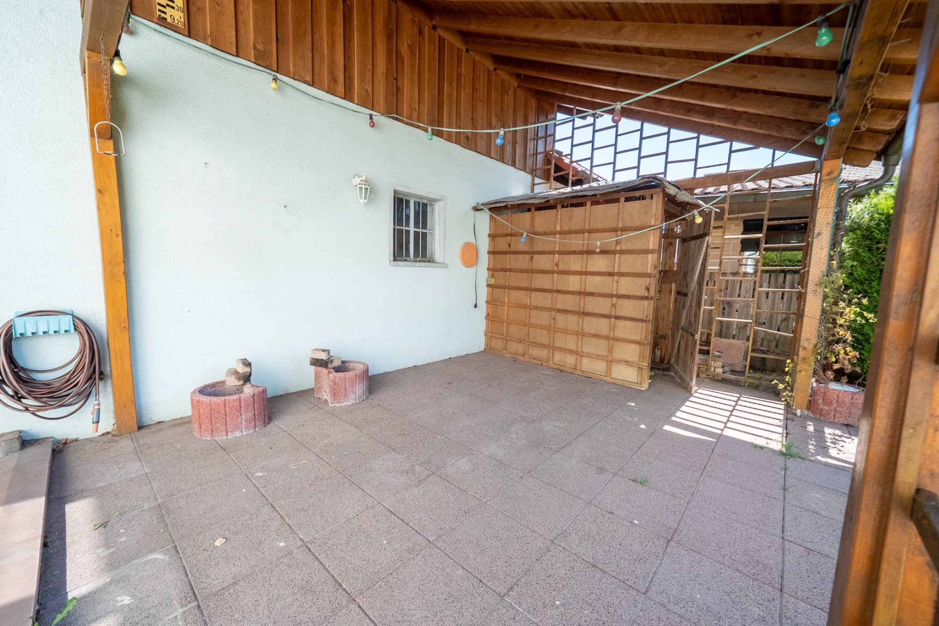 Überdachte Terrasse mit Pflastersteinen, Holzwänden und bunten Lichterketten.