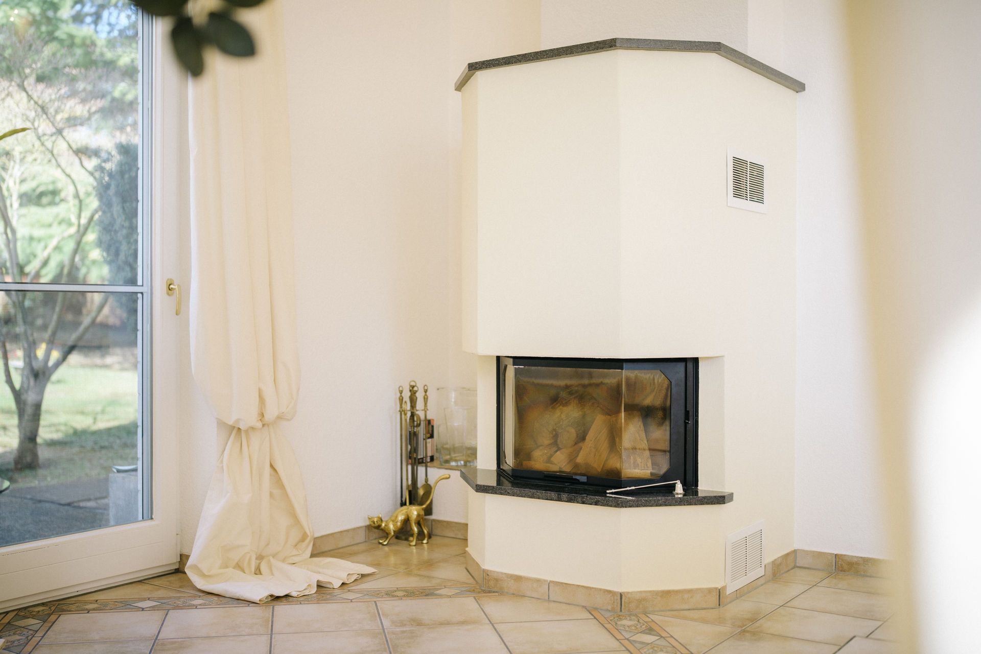 Heller Raum mit Kamin in der Ecke, bodentiefem Fenster und Vorhängen.