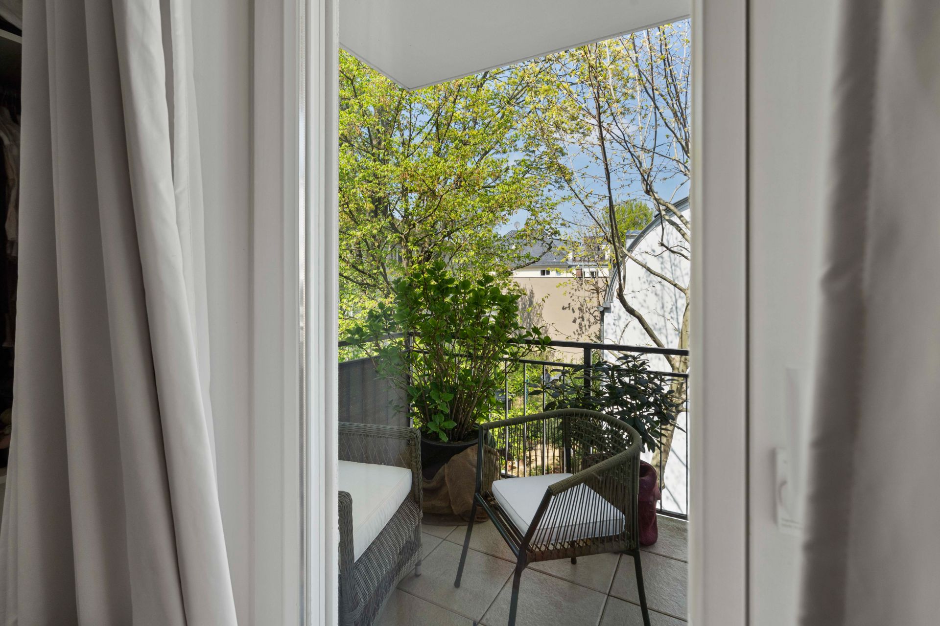 Blick durch Glastür auf einen Balkon mit zwei Stühlen, Pflanzen und Geländer.