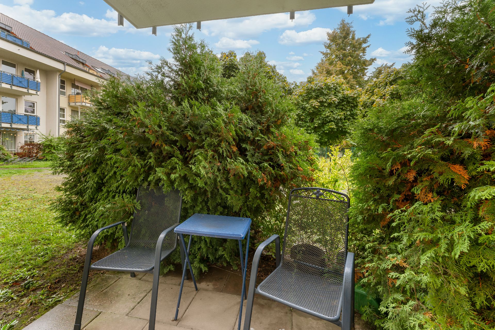 Zwei Metallstühle und ein kleiner Tisch stehen auf einer Terrasse mit Blick ins Grüne.