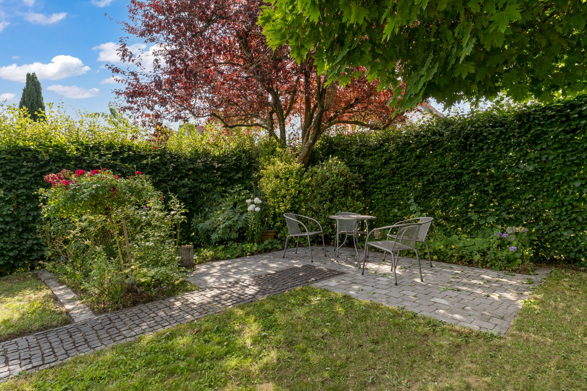 Kleiner Garten mit Terrasse, zwei Stühlen, Tisch und Blumenbeet, umgeben von Hecken und Bäumen.