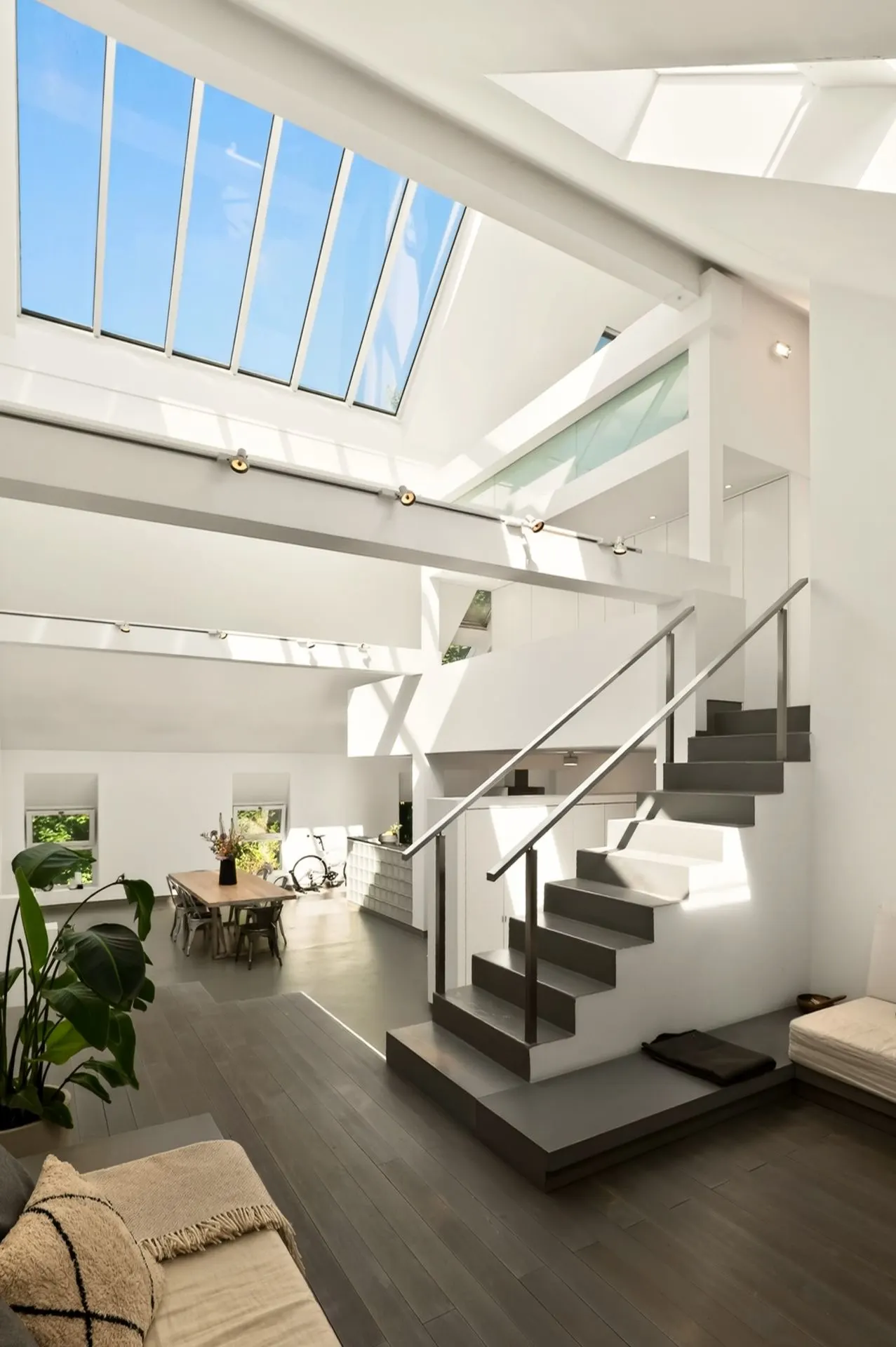 Helles Wohnzimmer mit großer Glasdecke, moderner Treppe und Esstisch, viele Pflanzen.