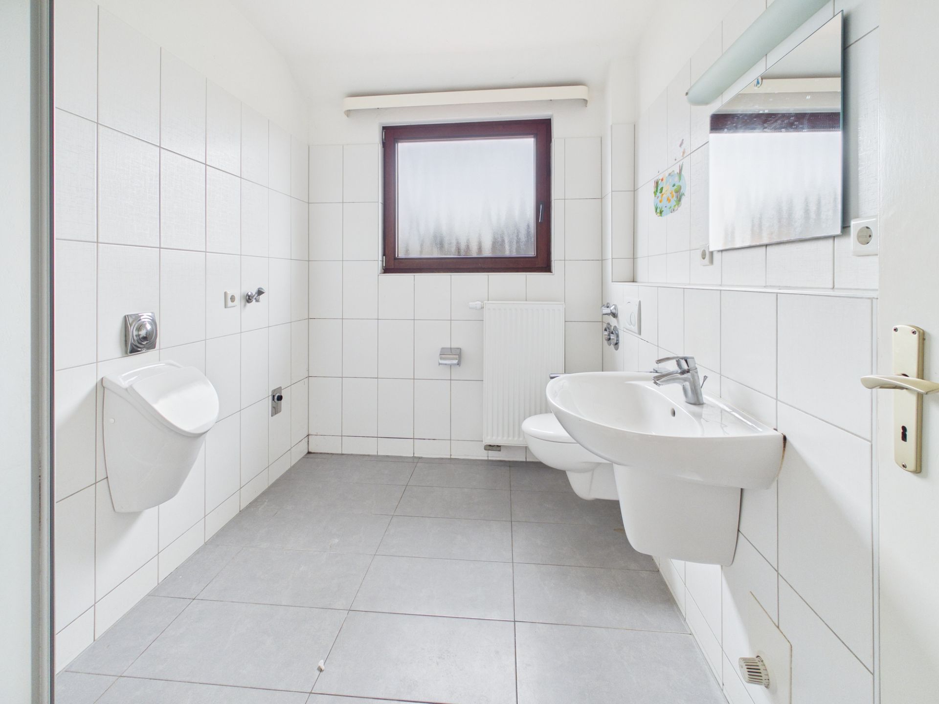 Helles Badezimmer mit weiß gefliesten Wänden, Urinal, Wandwaschbecken und Fenster.