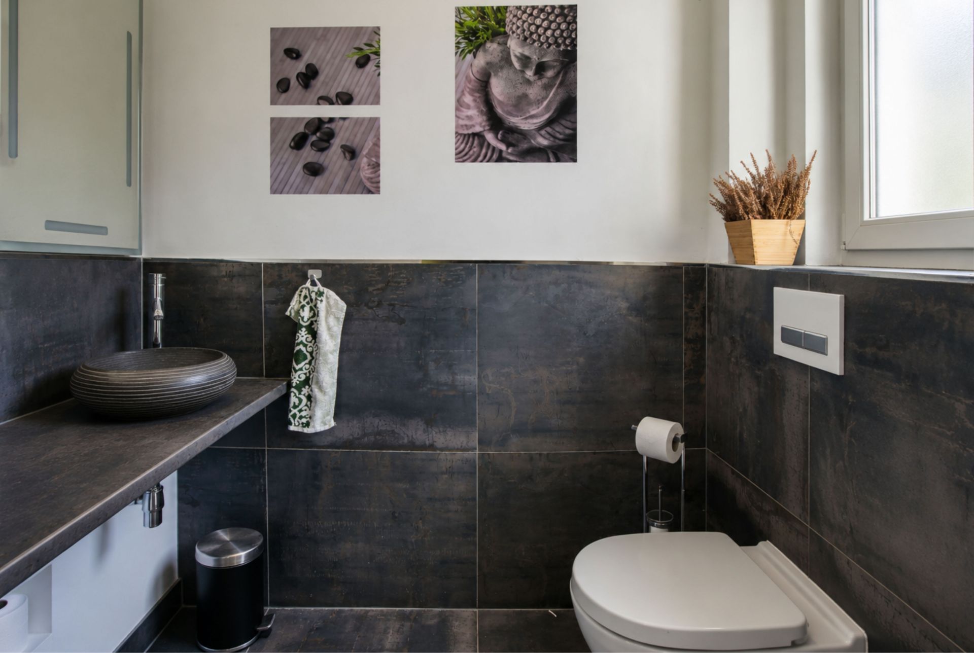 Modernes Badezimmer mit dunklen Fliesen, Waschbecken, Toilette, Fenster und Wandbildern.