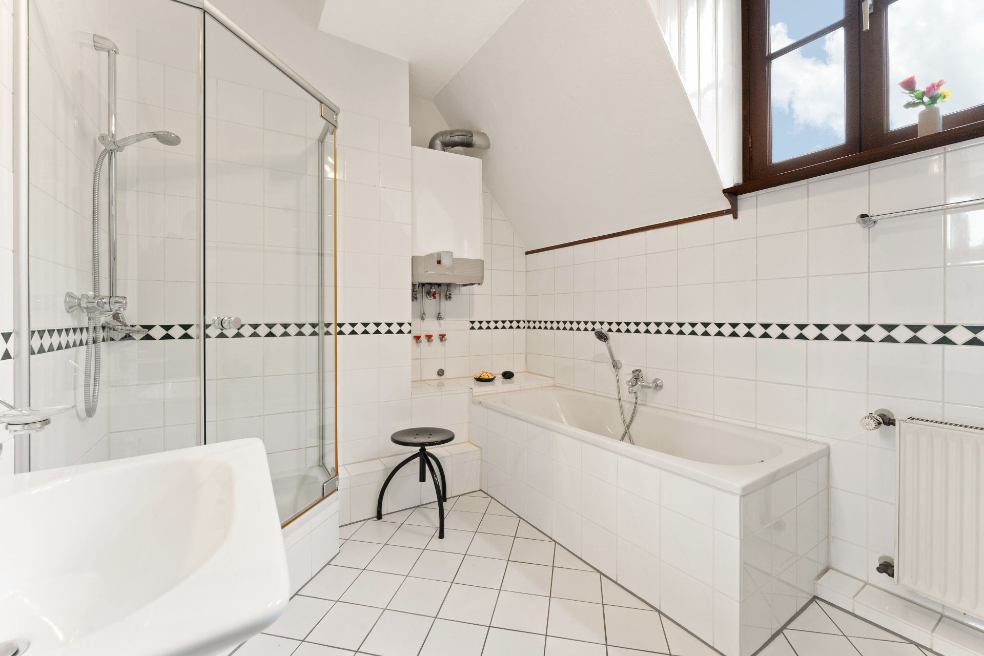Helles Bad mit Dusche, Badewanne, Waschbecken, Hocker und Fenster mit Blumen.