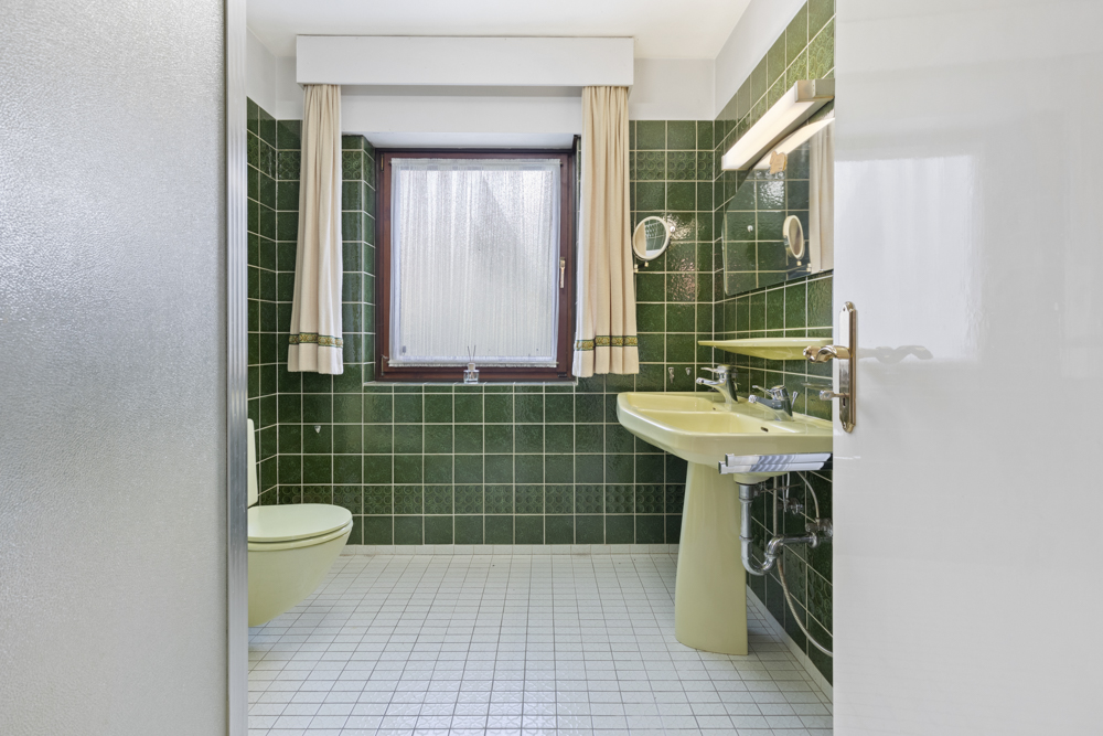Badezimmer mit grünen Fliesen, grünem WC, Waschbecken und Fenster mit Vorhängen.