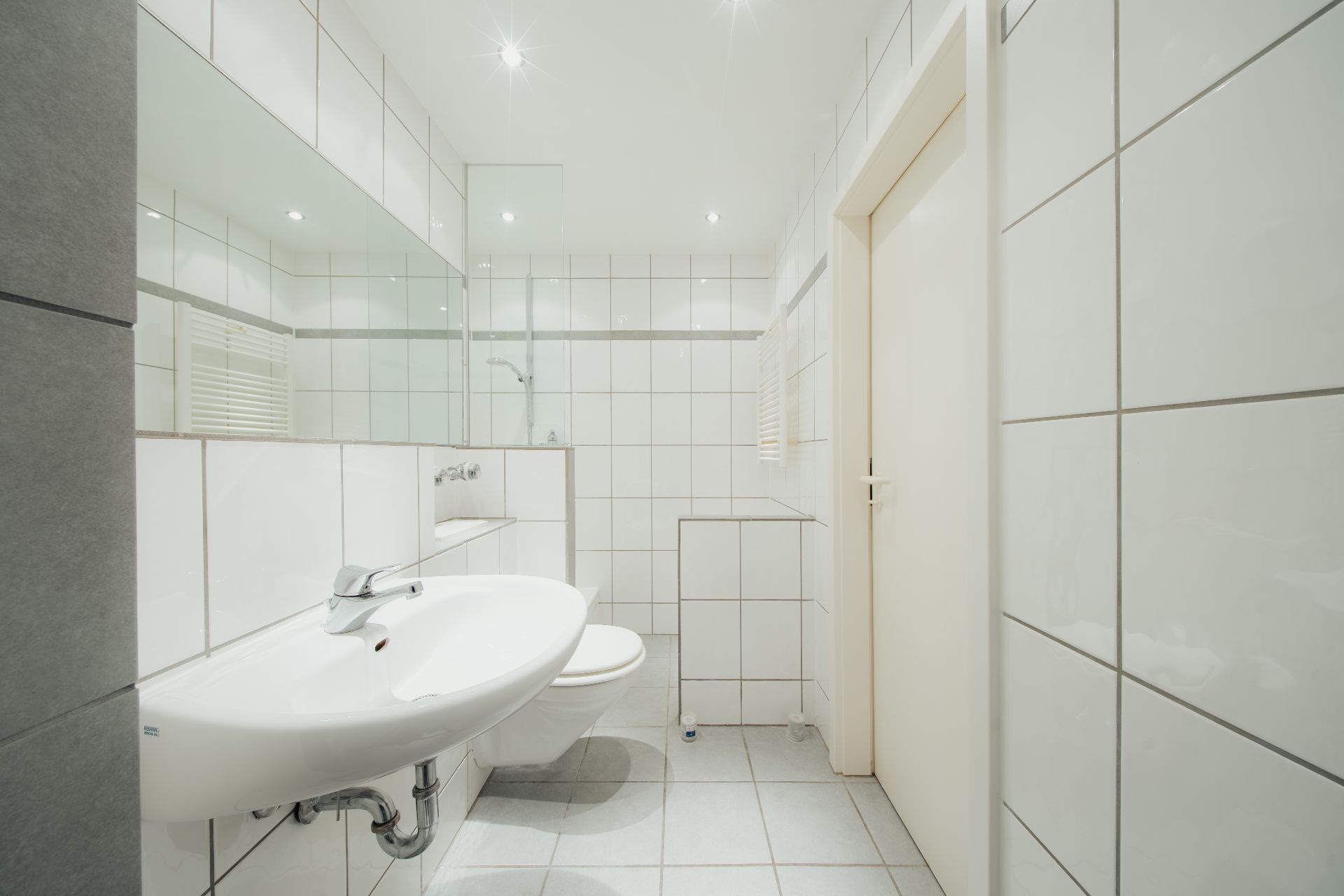 Helles Badezimmer mit weißen Fliesen, Waschbecken, WC und Spiegel.