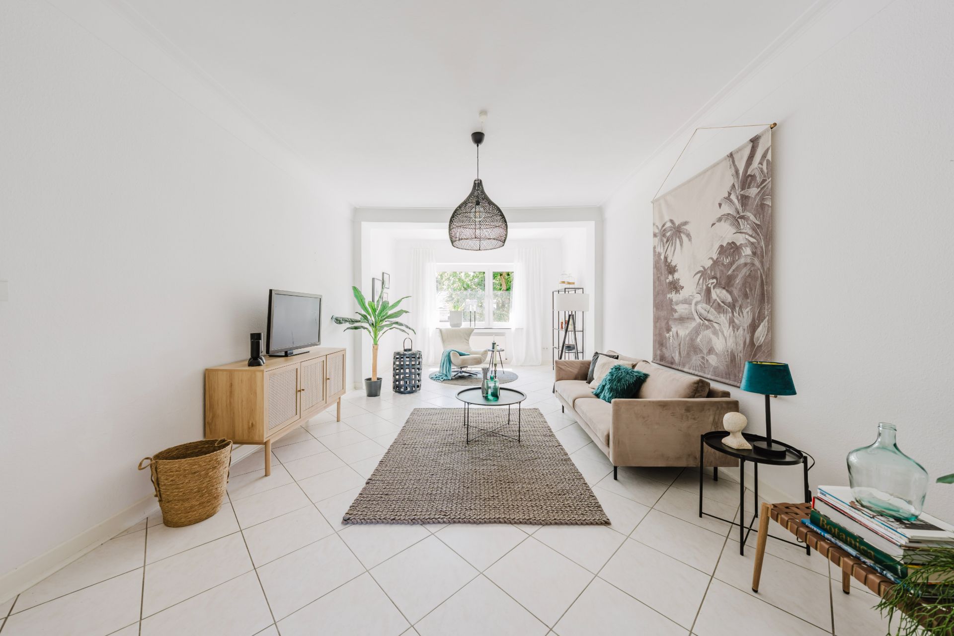 Helles Wohnzimmer mit Sofa, Fernseher, Teppich und Pflanzen, großes Fenster im Hintergrund.