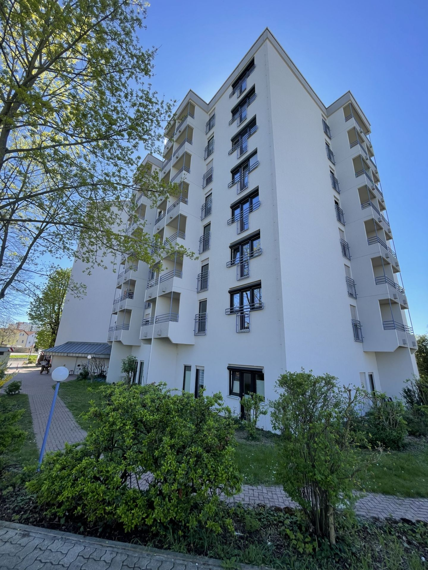 Mehrstöckiges Wohngebäude mit Balkonen, grünem Garten und Bäumen im Vordergrund.