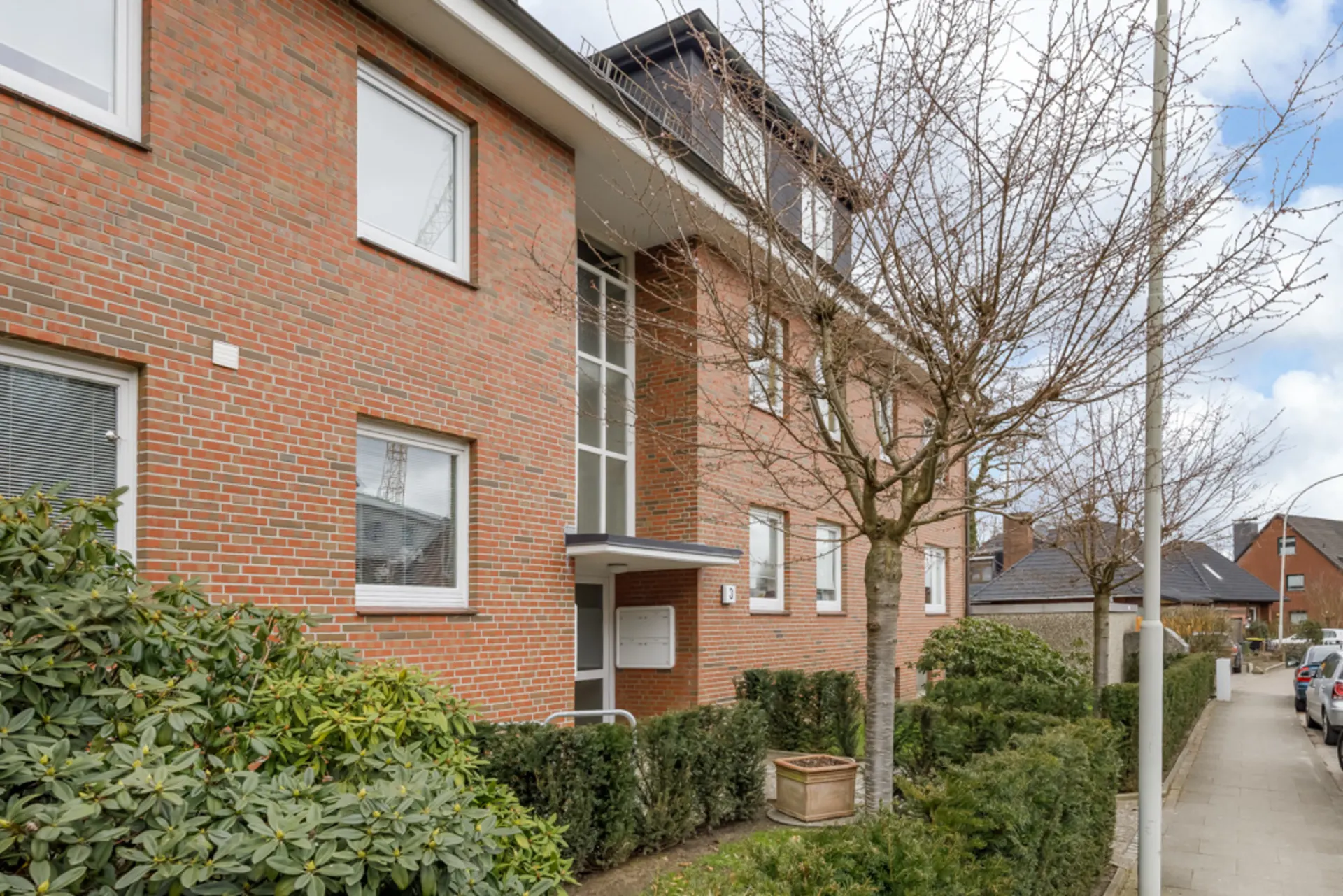 Mehrfamilienhaus aus rotem Backstein mit mehreren Fenstern, Bäumen und Gehweg davor.