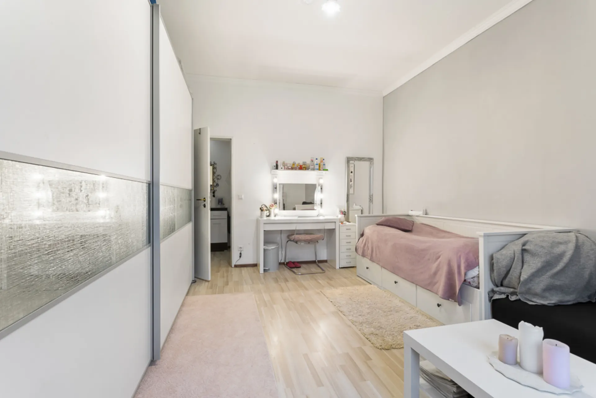 Helles Schlafzimmer mit Einzelbett, Schminktisch, großem Schrank und kleinem Tisch mit Kerzen.
