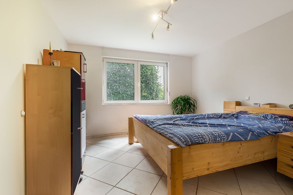 Schlafzimmer mit Holzbett, Schrank, großem Fenster und Pflanze auf Fliesenboden.