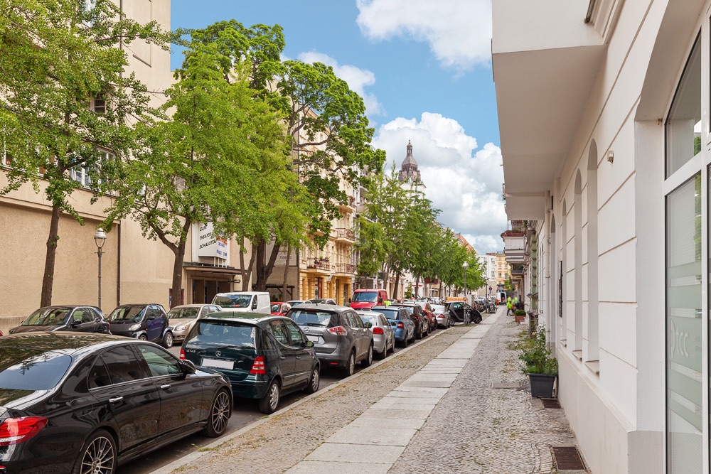 Wohnstraße mit geparkten Autos, Bäumen und mehrstöckigen Häusern bei Tageslicht.