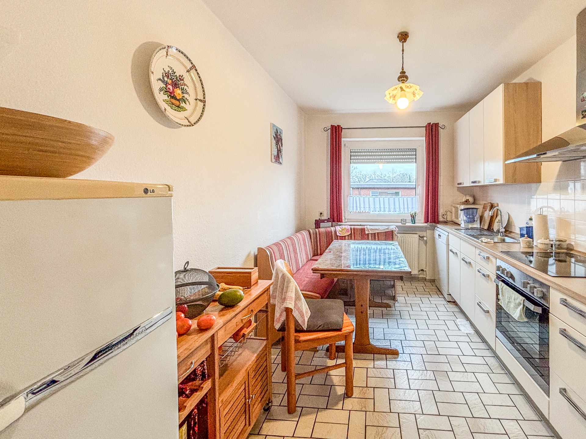 Helle Küche mit Esstisch, Eckbank, Kühlschrank, Herd und großem Fenster mit roten Vorhängen.