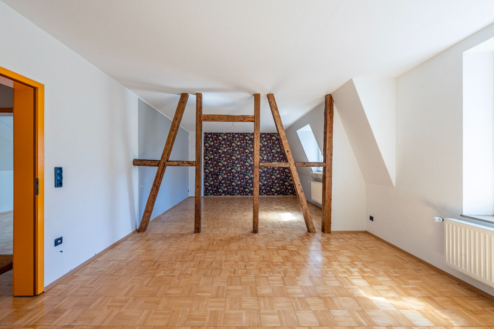 Heller Raum mit Holzstützen, Parkettboden und floral gemusterter Tapete an der Rückwand.