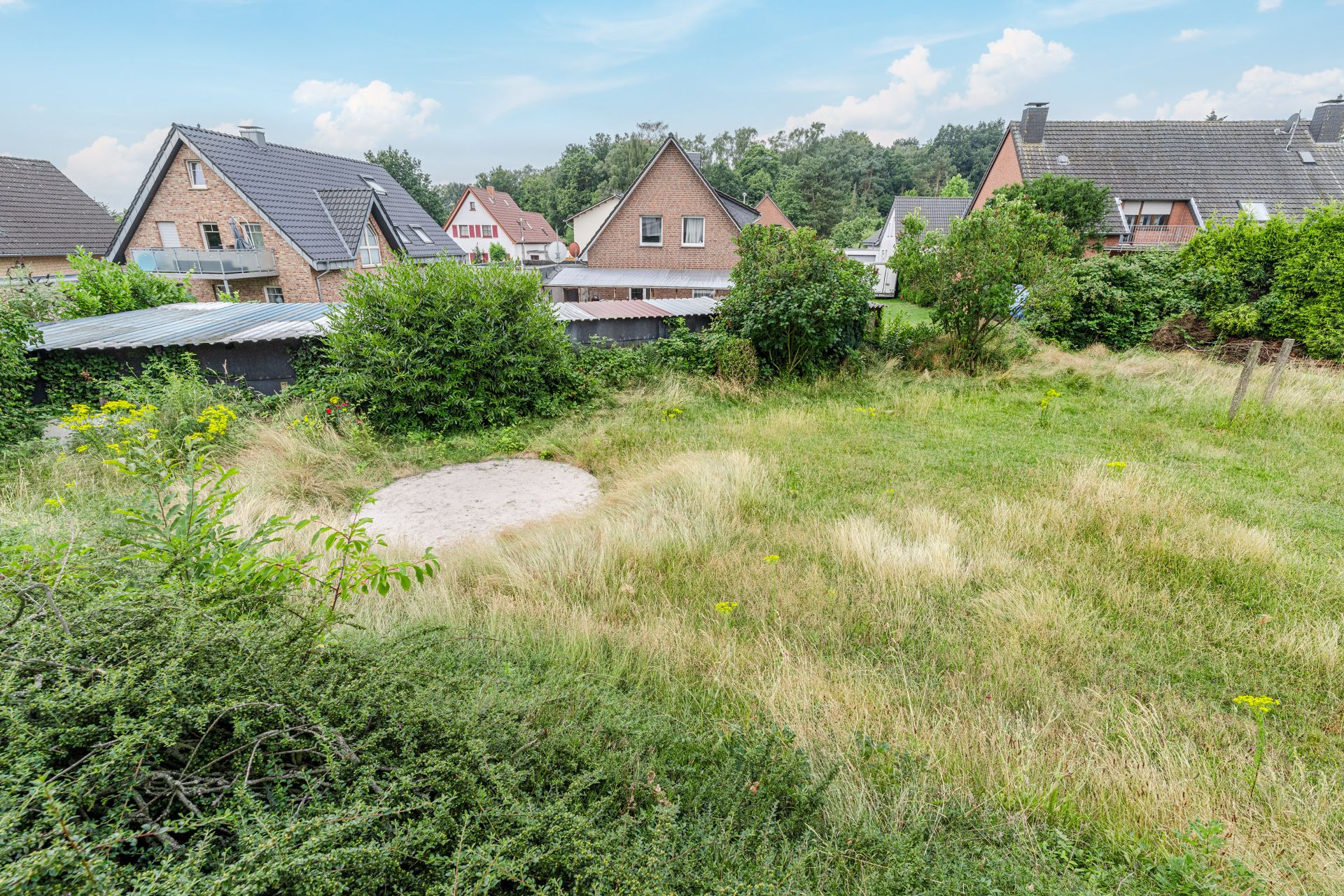 Garten mit hohem Gras, Sträuchern und einer Sandfläche, umgeben von Wohnhäusern.
