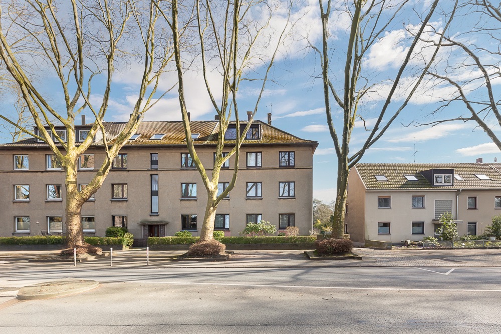 Zwei mehrstöckige Wohnhäuser mit vielen Fenstern und Bäumen davor an einer Straße.