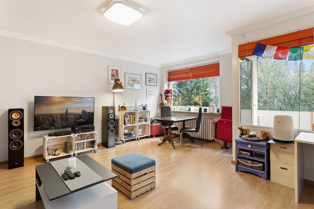 Helles Wohnzimmer mit Fernseher, Couchtisch, Schreibtisch und großen Fenstern mit Blick ins Grüne.