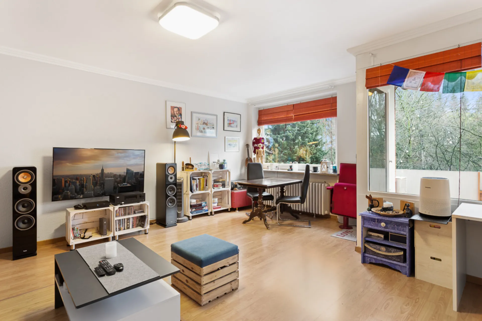 Helles Wohnzimmer mit Fernseher, Couchtisch, Schreibtisch und großen Fenstern mit Blick ins Grüne.