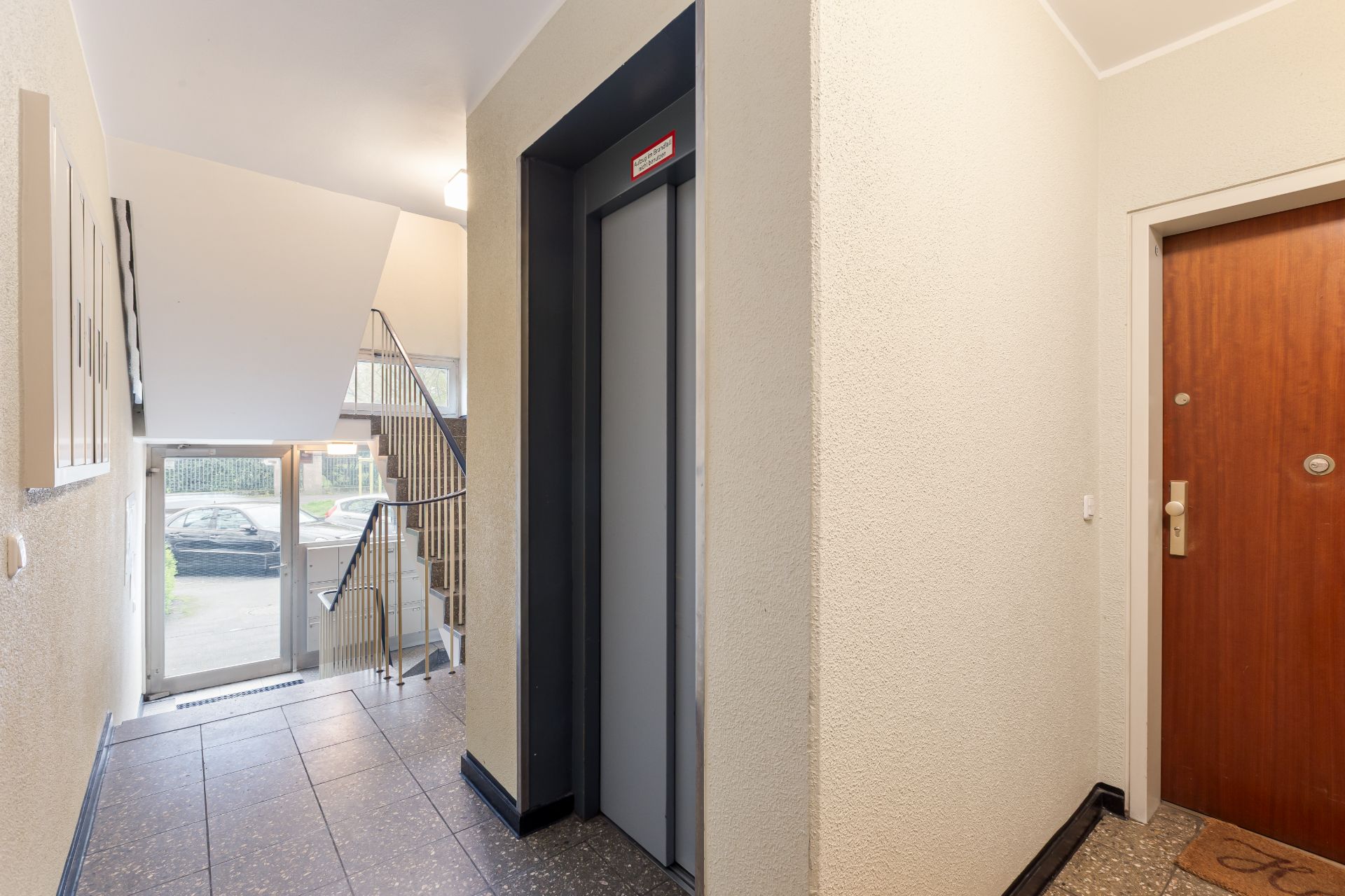 Hausflur mit Aufzug, Treppenhaus, Briefkästen und brauner Wohnungstür.