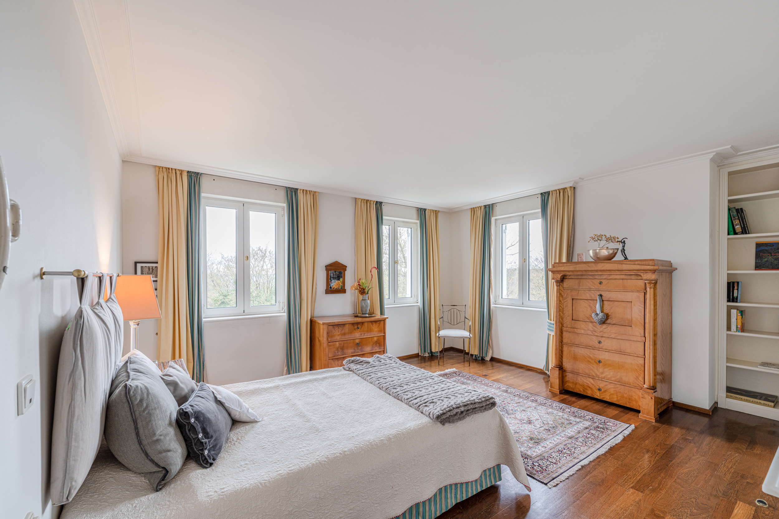 Helles Schlafzimmer mit Doppelbett, Holzboden, Schrank, Teppich und mehreren Fenstern mit Vorhängen.