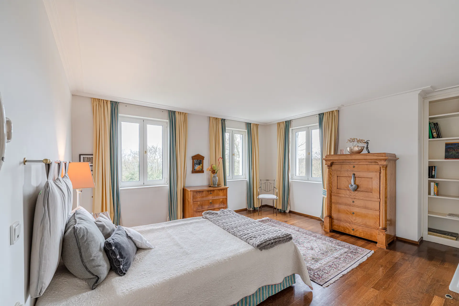 Helles Schlafzimmer mit Doppelbett, Holzboden, Schrank, Teppich und mehreren Fenstern mit Vorhängen.