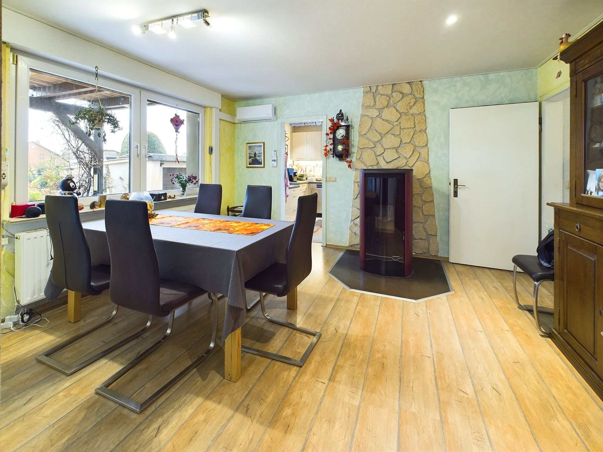Esszimmer mit Holzboden, Esstisch, sechs Stühlen, Kaminofen und großen Fenstern.
