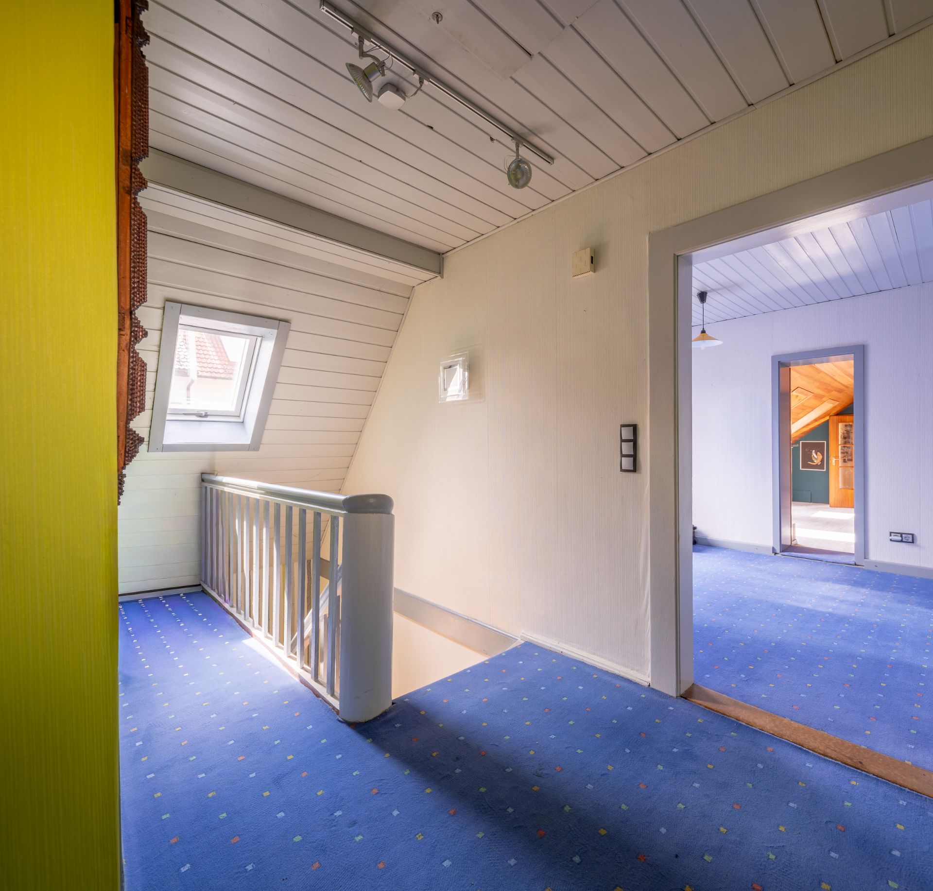 Obergeschossflur mit blauem Teppich, Treppengeländer und Dachfenster, angrenzender Raum sichtbar.