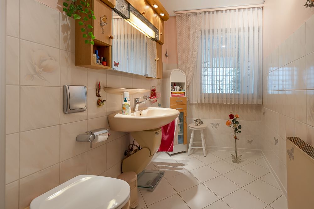 Helles Badezimmer mit Waschbecken, Spiegel, Toilette und Fliesen, dekoriert mit Pflanzen und Gardinen.