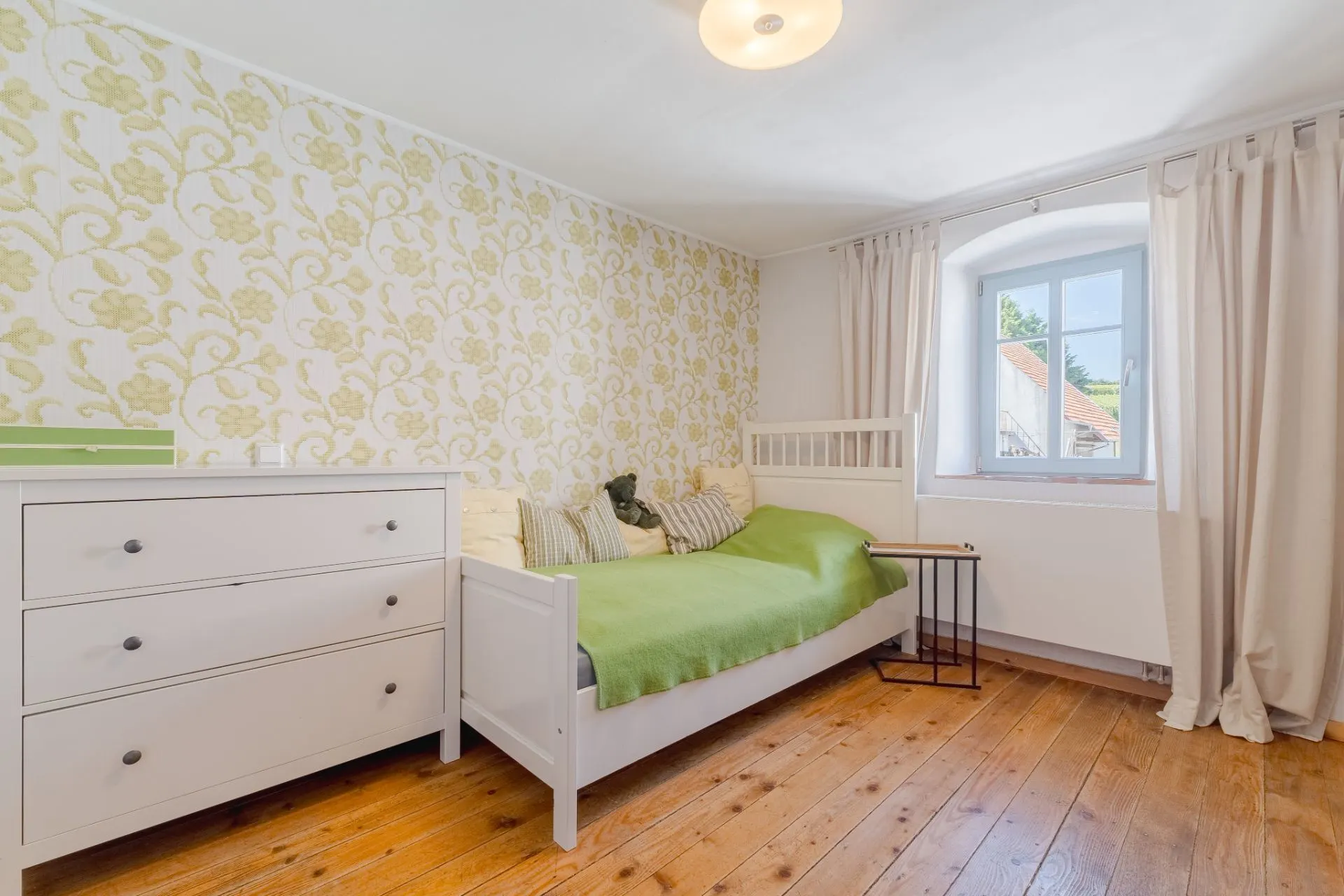 Helles Schlafzimmer mit Holzboden, Einzelbett, Kommode und Fenster mit Vorhängen.