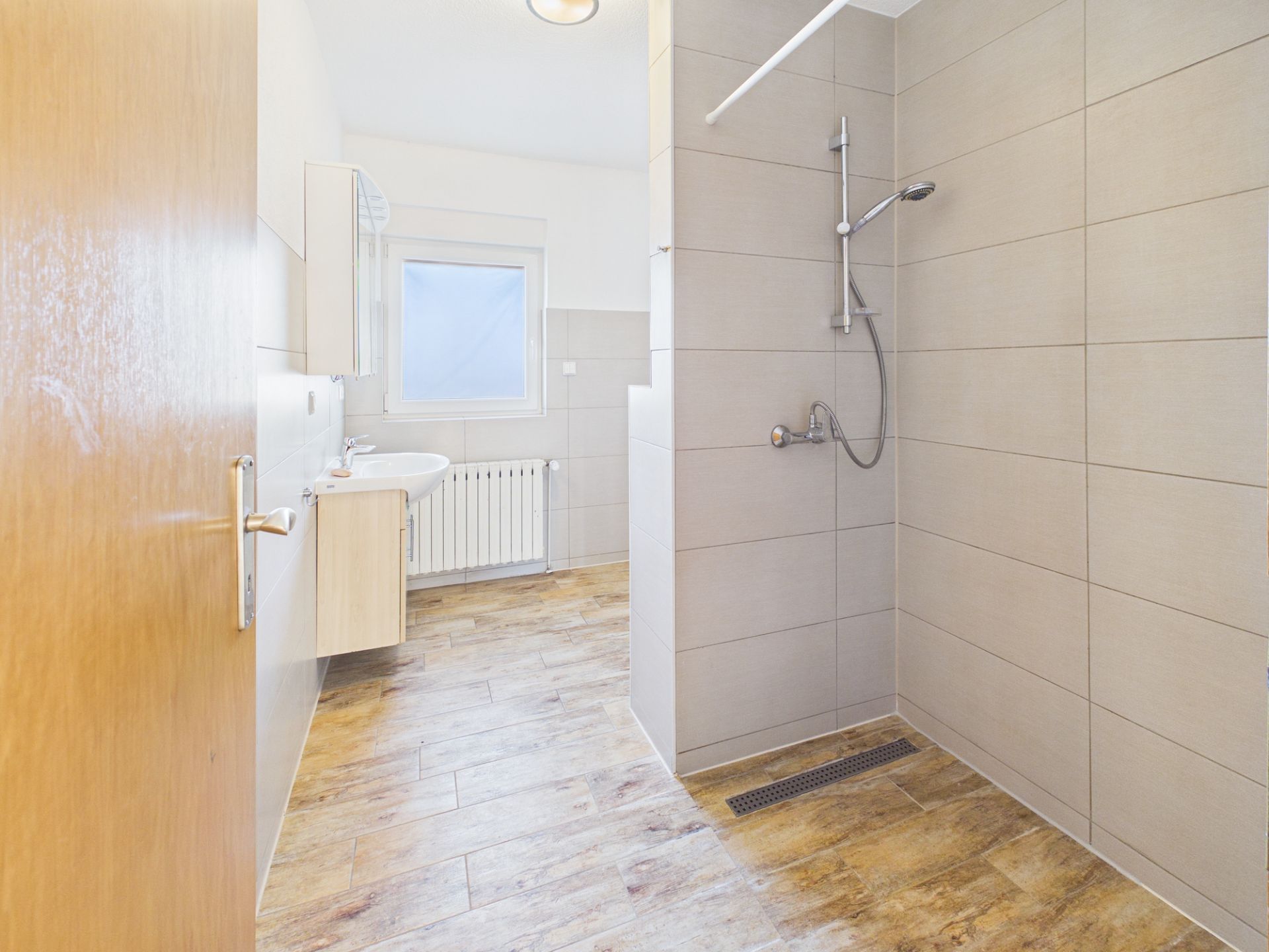 Modernes Badezimmer mit offener Dusche, Waschbecken, Fenster und Holzoptik-Fliesenboden.