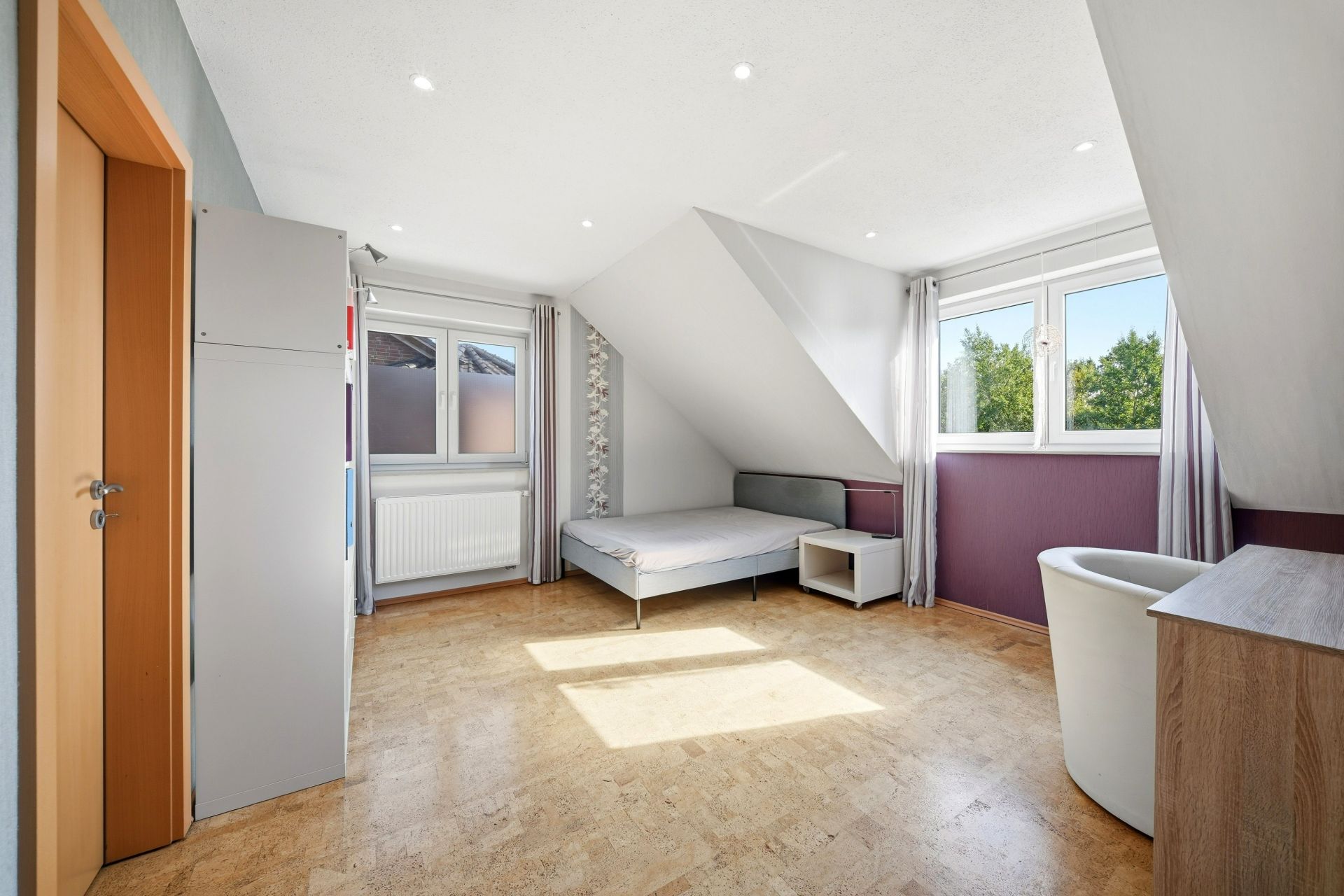 Helles Schlafzimmer mit Bett, Schrank, Sessel und zwei Fenstern im Dachgeschoss.