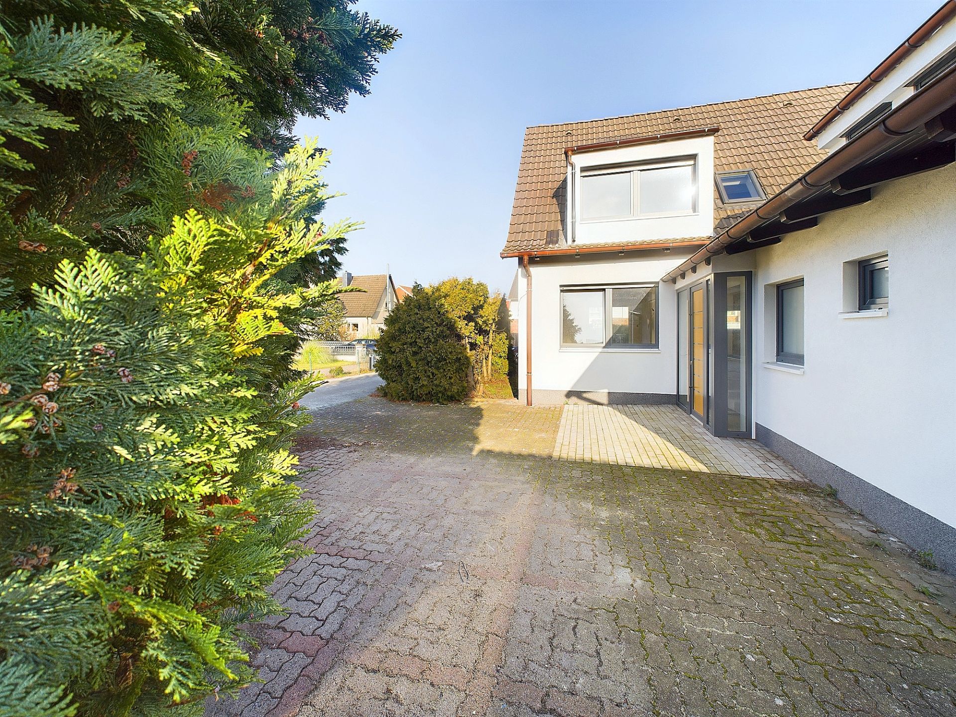 Gepflasterter Hauseingang mit Fenstern, Strauch und Baum im Vordergrund.