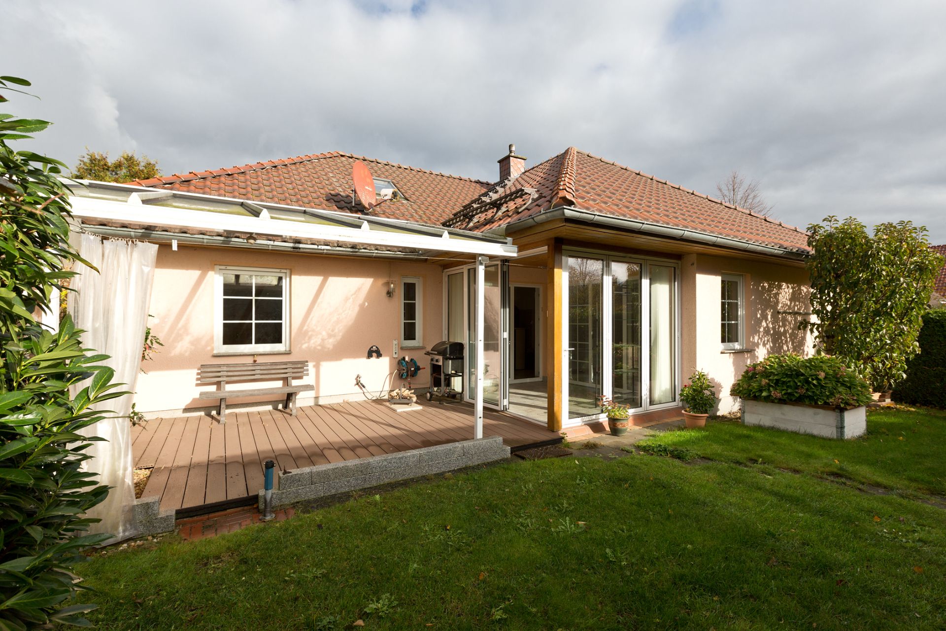 Haus mit Terrasse, überdachtem Sitzbereich und Garten, große Fenster und Pflanzenbeete sichtbar.