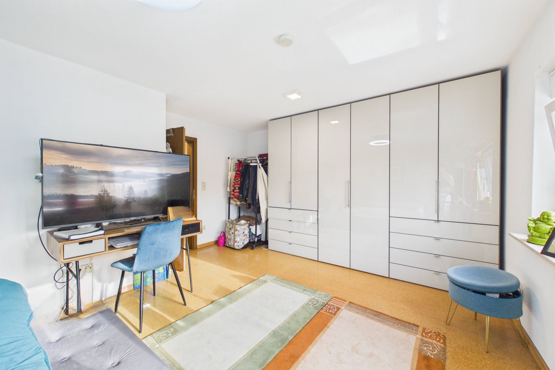 Helles Zimmer mit großem Kleiderschrank, Schreibtisch, TV, Stuhl, Spiegel und Teppich.