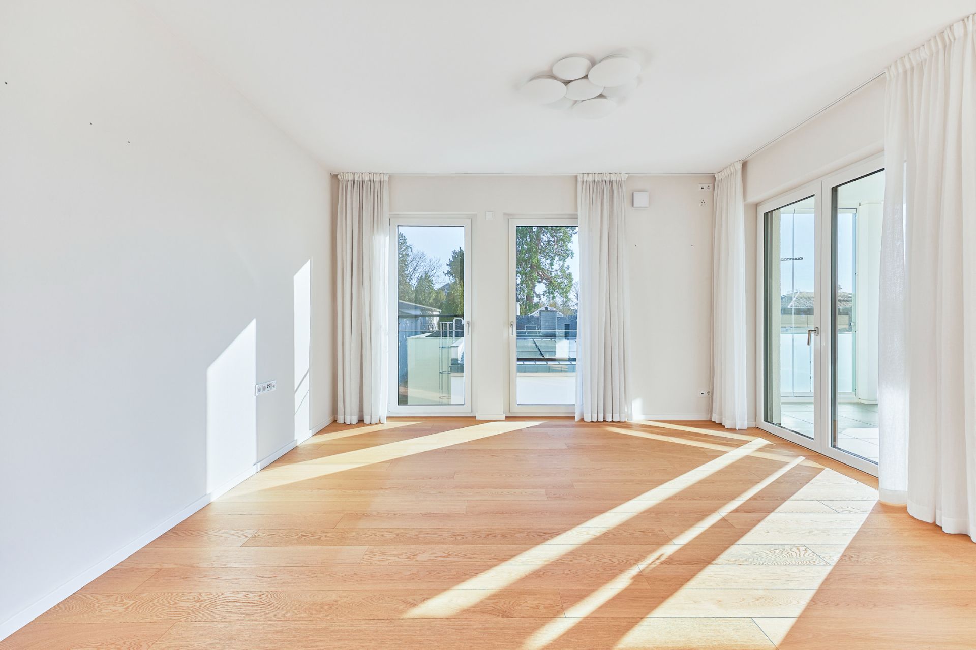 Helles Wohnzimmer mit Holzfußboden, großen Fenstern und weißen Vorhängen.