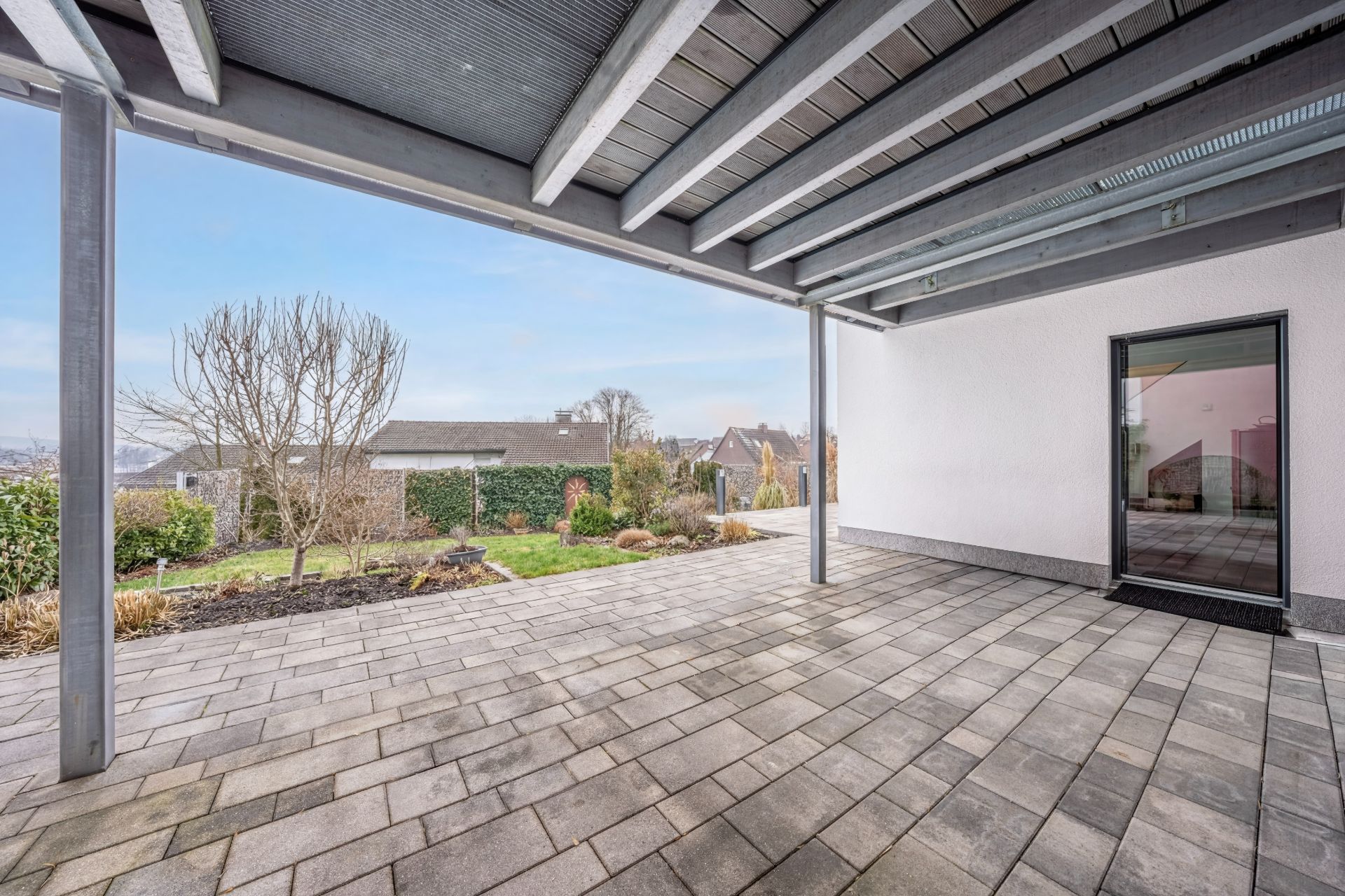 Überdachte Terrasse mit Steinboden, Blick auf Garten mit Bäumen und Sträuchern.