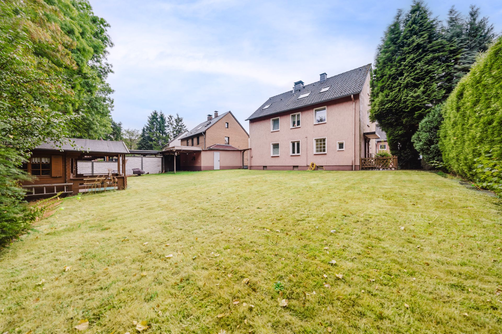 Großer Garten mit Rasenfläche, einem Wohnhaus und einer überdachten Terrasse links.