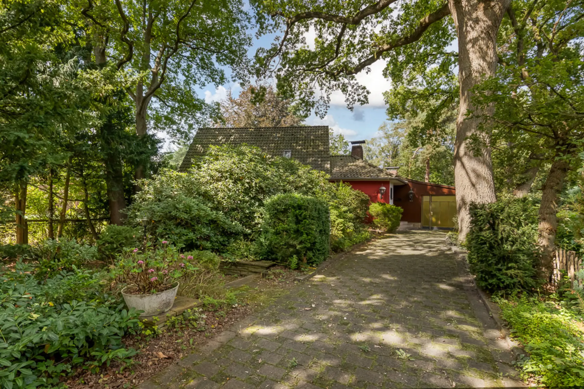 Haus mit Ziegeldach, gepflasterter Einfahrt und viel grünem Garten, von großen Bäumen umgeben.