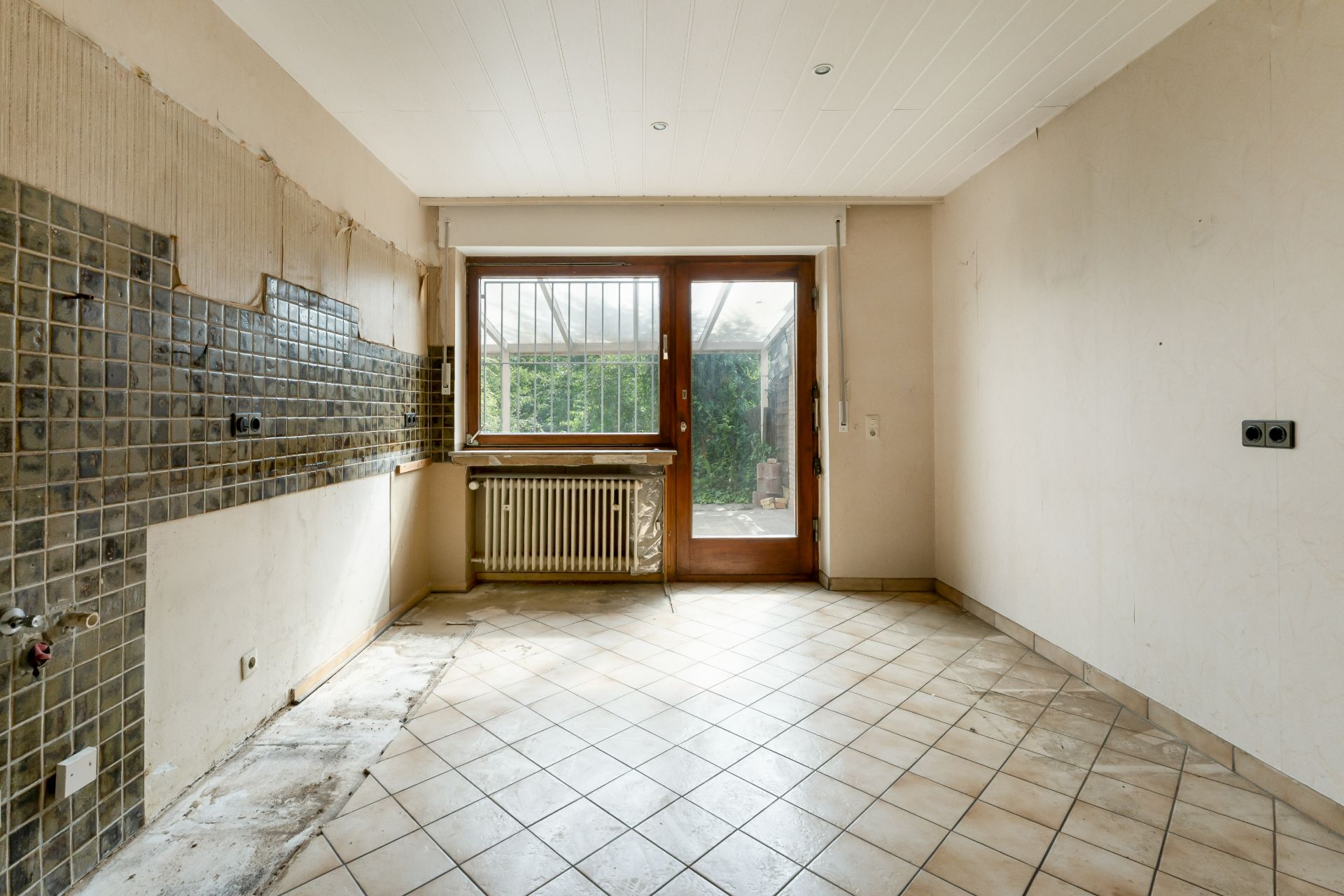 Leerer Raum mit Fliesenboden, braunen Wandfliesen, Fenster und Glastür zum Garten.