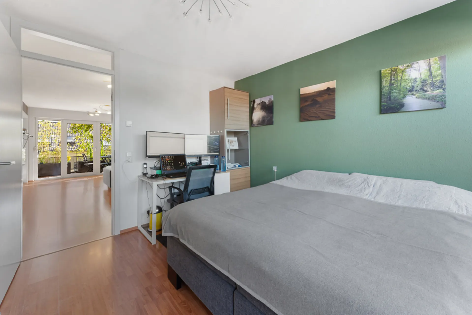 Schlafzimmer mit Doppelbett, Schreibtisch mit Monitoren und grüner Wand mit Bildern.