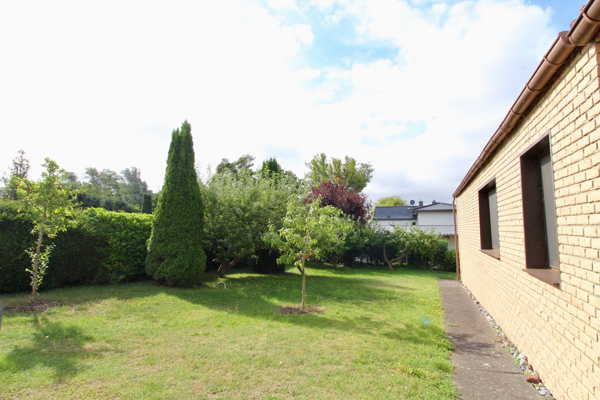 Garten mit Rasen, Bäumen und Sträuchern neben einem Gebäude mit gelber Ziegelwand.