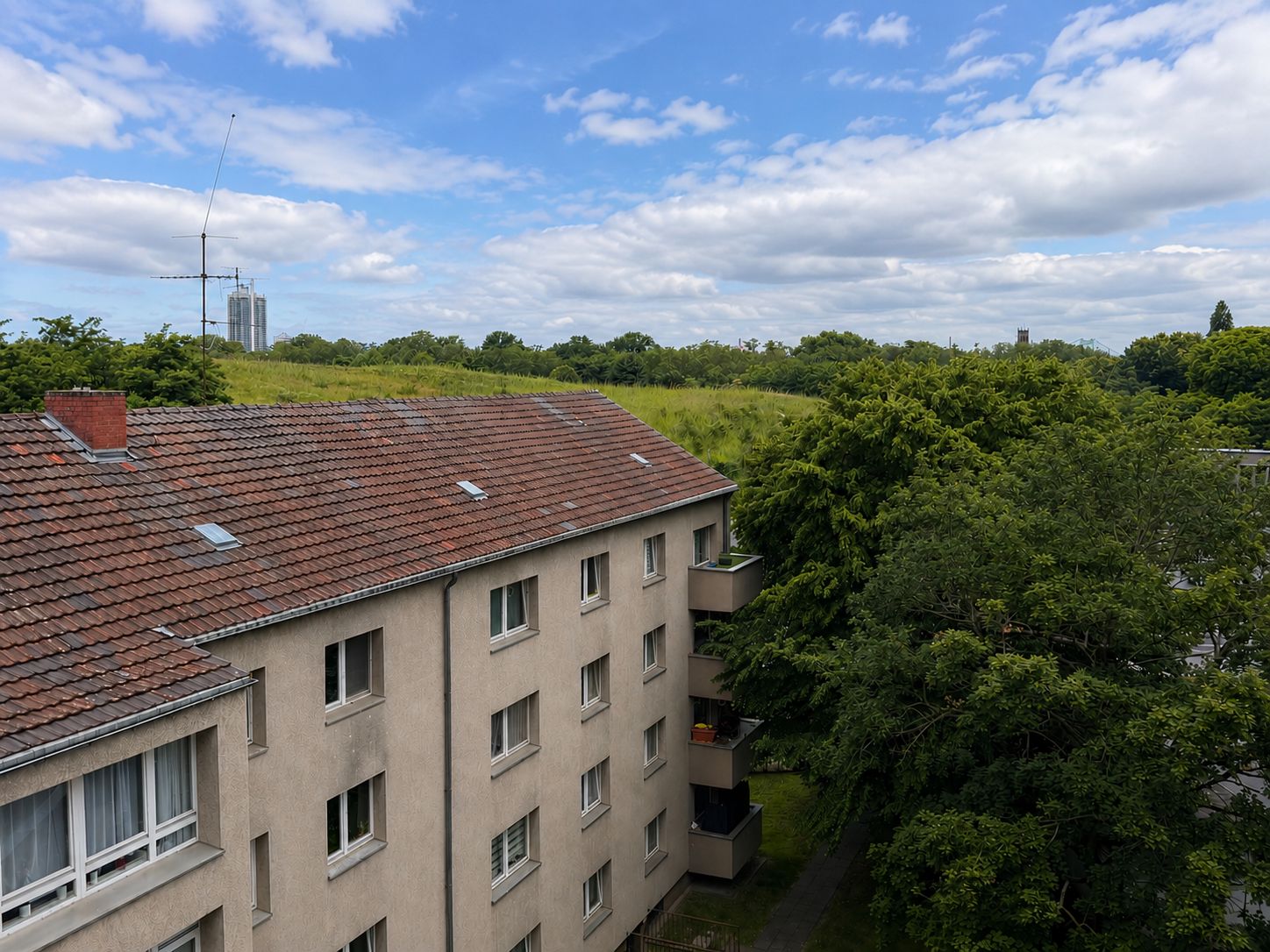 Mehrfamilienhaus mit rotem Dach und mehreren Fenstern, daneben Bäume und Wiese unter blauem Himmel.