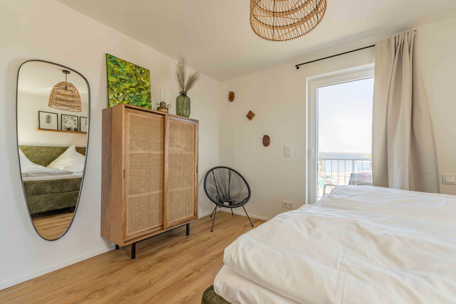 Helles Schlafzimmer mit Bett, Holzschrank, Spiegel, Stuhl und Fenster mit Vorhang.